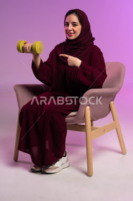 ممارسة الأنشطة الرياضية, بورتريه لسيدة عربية خليجية سعودية محجبة مبتسمة ترتدي العباءة الملونة تنظر إلى الكاميرا تحمل بيدها الاوزان الثقيلة الحديدية ( الدمبلز) تشير إليها, اتباع أسلوب حياة صحي, مفهوم اللياقة والليونة والرشاقة البدنية, خلفية بنفسجية