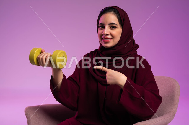 هواية ممارسة الأنشطة الرياضية, بورتريه مقرب لسيدة عربية خليجية سعودية محجبة مبتسمة ترتدي العباءة الملونة تنظر إلى الكاميرا تحمل بيدها الاوزان الثقيلة الحديدية ( الدمبلز) تشير إليها, اتباع أسلوب حياة صحي, مفهوم اللياقة والليونة والرشاقة البدنية, خلفية بنفسجية