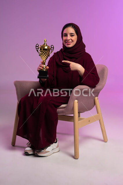 إيماءات السرور بفوز الفريق المفضل, مشاركة النساء السعوديات في الأنشطة الرياضية، تشجيع الأنديه والمنتخبات، بورتريه لشابة عربية سعودية خليجية مبتسمة ترتدي العباءه الملونة تجلس على كرسي مريح تنظر إلى الكاميرا تشير بإصبع السبابة إلى الكأس، خلفية بنفسجية