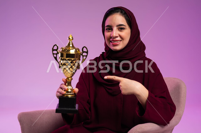 مشاركة النساء السعوديات في الأنشطة الرياضية، تشجيع الأنديه والمنتخبات، بورتريه مقرب لشابة عربية سعودية خليجية مبتسمة ترتدي العباءه الملونة تجلس على كرسي مريح تنظر إلى الكاميرا تشير بإصبع السبابة إلى الكأس، إيماءات السرور بفوز الفريق المفضل, خلفية بنفسجية