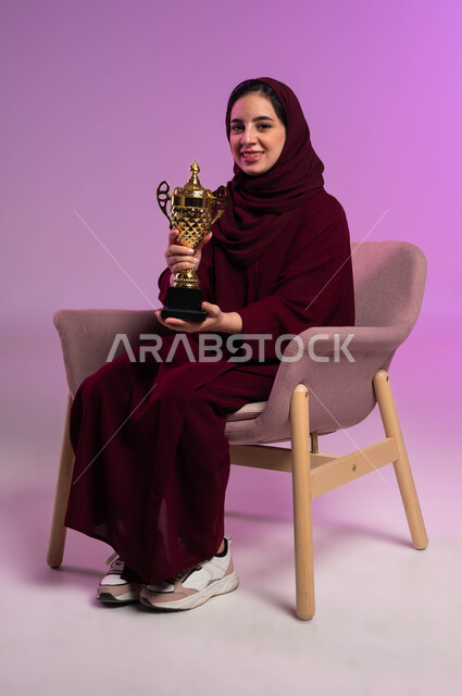 أنشطة رياضية ترفيهية, متابعة ومشاهدة المباريات، تشجيع الأنديه والمنتخبات، بورتريه لشابة عربية سعودية خليجية مبتسمة ترتدي العباءه الملونة تجلس على كرسي مريح تنظر إلى الكاميرا ترفع يدها تحمل الكأس، خلفية بنفسجية