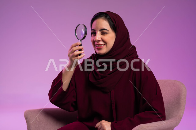 التدقيق والتركيز وتكبير الاشياء, الفحص والإستكشاف والقراءة، بورتريه مقرب لشابة عربية خليجية سعودية محجبة ترتدي العباءة الملونة تجلس على كرسي مريح تنظر من خلال العدسة المكبرة، أدوات بصرية للرؤية عن قرب، مفهوم توضيح الصورة، خلفية بنفسجية