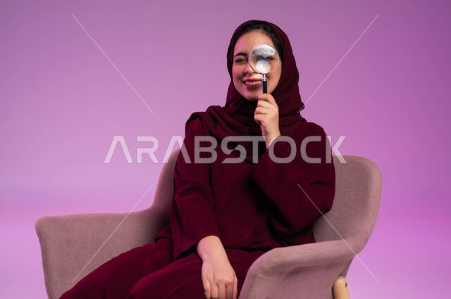 الفحص والإستكشاف والقراءة، مفهوم توضيح الصورة، بورتريه مقرب لشابة عربية خليجية سعودية محجبة ترتدي العباءة الملونة تجلس على كرسي مريح تنظر من خلال العدسة المكبرة، التدقيق والتركيز وتكبير الاشياء, أدوات بصرية للرؤية عن قرب، خلفية بنفسجية