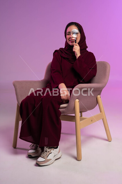 مفهوم توضيح الصورة، بورتريه لشابة عربية خليجية سعودية محجبة ترتدي العباءة الملونة تجلس على كرسي مريح تنظر من خلال العدسة المكبرة، الفحص والإستكشاف والقراءة، التدقيق والتركيز وتكبير الاشياء, أدوات بصرية للرؤية عن قرب، خلفية بنفسجية