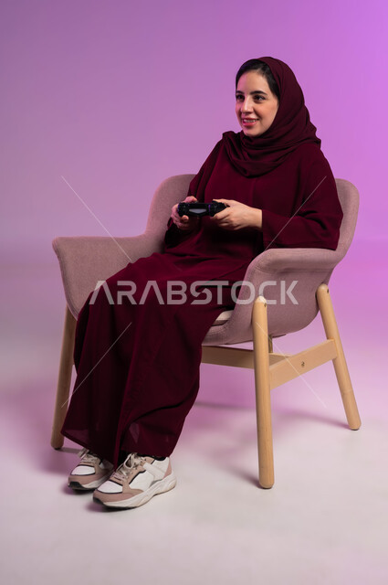 قضاء إجازة صيفية ممتعة، إيماءات الاستمتاع والاندماج والتفاعل, تقنيات الكترونية حديثة, بورتريه لإمرأة عربية سعودية خليجية تجلس على كرسي مريح تحمل بيدها عصا التحكم تلعب في ألعاب البلايستيشن الترفيهية، خلفية بنفسجية