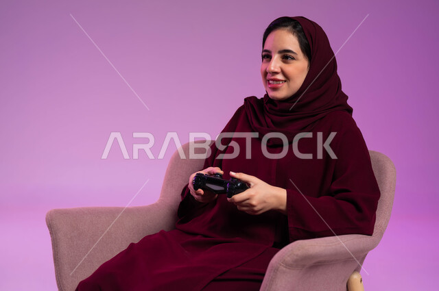 إيماءات الاستمتاع والاندماج والتفاعل, قضاء إجازة صيفية ممتعة، تقنيات الكترونية حديثة, بورتريه مقرب لإمرأة عربية سعودية خليجية تجلس على كرسي مريح تحمل بيدها عصا التحكم تلعب في ألعاب البلايستيشن الترفيهية، خلفية بنفسجية