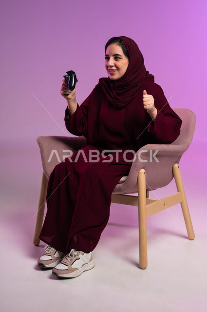 إيماءات الفوز والحماس, التقنيات الالكترونية الحديثة, مفهوم الاندماج والتفاعل باللعب في ألعاب البلايستيشن الترفيهية، بورتريه لإمرأة عربية سعودية خليجية تجلس على كرسي مريح تحمل بيدها عصا التحكم، قضاء أوقات ممتعة في الإجازة, خلفية بنفسجية