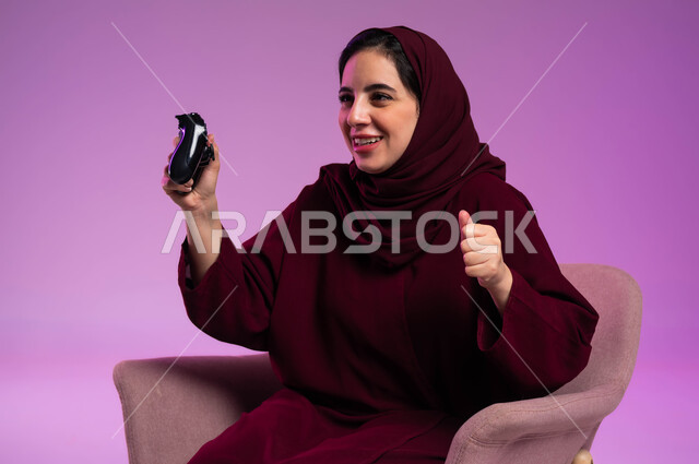 قضاء أوقات ممتعة في الإجازة, إيماءات الفوز والحماس, التقنيات الالكترونية الحديثة, مفهوم الاندماج والتفاعل باللعب في ألعاب البلايستيشن الترفيهية، بورتريه مقرب لإمرأة عربية سعودية خليجية تجلس على كرسي مريح تحمل بيدها عصا التحكم، خلفية بنفسجية