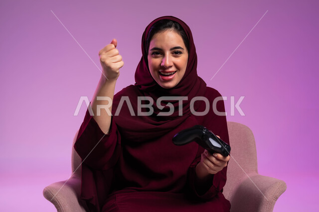 التفاعل والاندماج باللعب في ألعاب البلايستيشن الترفيهية، التقنيات الالكترونية الحديثة, بورتريه مقرب لإمرأة عربية سعودية خليجية تجلس على كرسي مريح تنظر إلى الكاميرا تحمل عصا التحكم ترفع يدها للأعلى بإيماءات الفوز والانتصار، خلفية بنفسجية