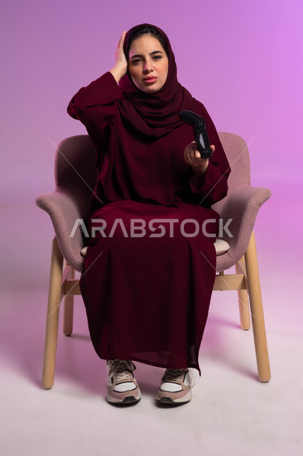 Modern electronic technologies, gestures of sadness and loss, spending free time playing recreational PlayStation games, a portrait of a Saudi Arabian Gulf woman sitting in a comfortable chair looking at the camera, holding a joystick and placing her hand on her head, purple background