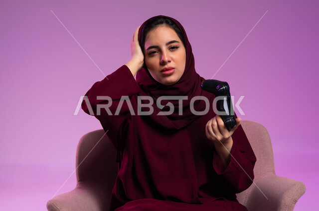 إيماءات الحزن والخسارة, قضاء أوقات الفراغ باللعب في ألعاب البلايستيشن الترفيهية، تقنيات الكترونية حديثة, بورتريه مقرب لإمرأة عربية سعودية خليجية تجلس على كرسي مريح تنظر إلى الكاميرا تحمل عصا التحكم تضع يدها على رأسها، خلفية بنفسجية