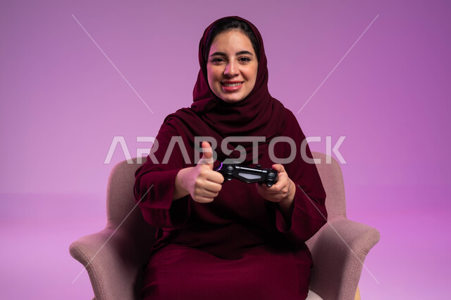 قضاء أوقات الفراغ باللعب في ألعاب البلايستيشن الترفيهية، إيماءات الإعجاب والرضا, تقنيات الكترونية حديثة, بورتريه مقرب لإمرأة عربية سعودية خليجية تجلس على كرسي مريح تنظر إلى الكاميرا تحمل بيدها عصا التحكم ترفع إصبع الإبهام نحو الأعلى، خلفية بنفسجية