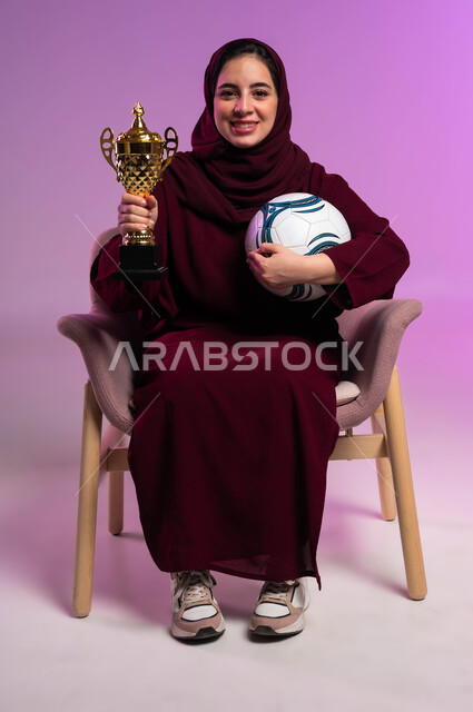 الأنشطة الرياضية الترفيهية والمباريات، النظر إلى الكاميرا بإيماءات السعادة، بورتريه مقرب لشابة عربية سعودية خليجية مبتسمة ترتدي العباءه الملونة تجلس على كرسي مريح  تحمل بيدها كرة القدم وكأس الفوز، تشجيع الأنديه والمنتخبات، خلفية بنفسجية