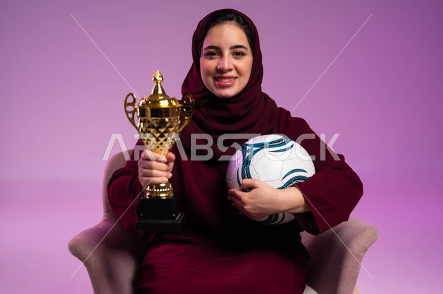 الشغف في مباريات كرة القدم، النظر إلى الكاميرا بإيماءات السعادة، بورتريه مقرب لشابة عربية سعودية خليجية مبتسمة ترتدي العباءه الملونة تجلس على كرسي مريح تحمل بيدها الكرة وكأس الفوز، تشجيع الأنديه والمنتخبات، خلفية بنفسجية