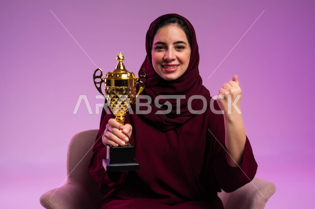 تشجيع الأنديه والمنتخبات، الشغف في مباريات كرة القدم والرياضات الترفيهية، بورتريه مقرب لشابة عربية سعودية خليجية مبتسمة ترتدي العباءه الملونة تجلس على كرسي مريح تنظر إلى الكاميرا تحمل الكأس ترفع يدها بإيماءات الفوز، خلفية بنفسجية