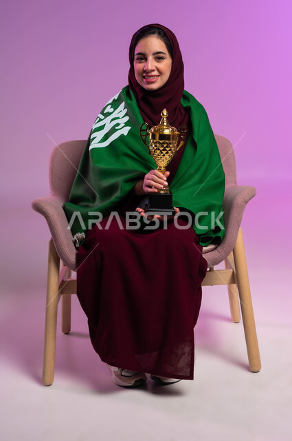 الشغف في مباريات كرة القدم والأنشطة الرياضية، النظر إلى الكاميرا بإيماءات السعادة، بورتريه لشابة عربية سعودية خليجية مبتسمة ترتدي العباءه الملونة تجلس على كرسي مريح تضع على ظهرها علم المملكة تحمل بيدها كأس الفوز، تشجيع الأنديه والمنتخبات، خلفية بنفسجية