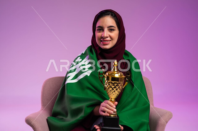 مشاركة النساء السعوديات في المباريات والأنشطة الرياضية, تشجيع الأنديه والمنتخبات، بورتريه مقرب لشابة عربية سعودية خليجية مبتسمة ترتدي العباءه الملونة تجلس على كرسي مريح تضع على ظهرها علم المملكة تنظر إلى الكاميرا تحمل بيدها الكأس، خلفية بنفسجية