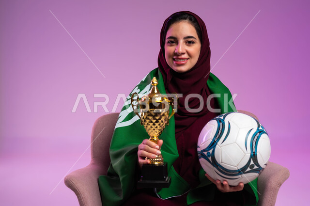 الشغف في مباريات كرة القدم، النظر إلى الكاميرا بإيماءات السعادة، بورتريه مقرب لشابة عربية سعودية خليجية مبتسمة ترتدي العباءه الملونة تجلس على كرسي مريح تضع على ظهرها علم المملكة تحمل بيدها الكرة وكأس الفوز، تشجيع الأنديه والمنتخبات، خلفية بنفسجية