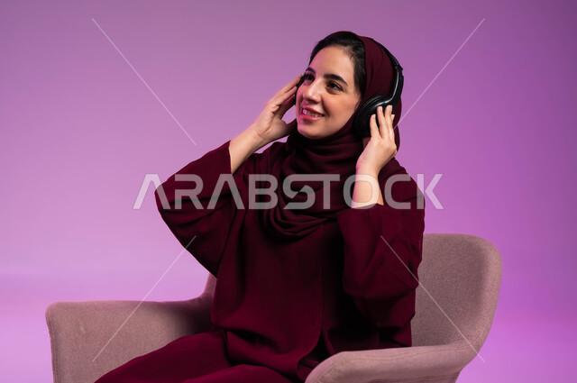 Enjoying leisure time, the concept of listening and listening, using headphone techniques, a close-up portrait of a smiling Saudi Gulf Arab woman, veiled, wearing a colorful abaya, wearing audio headphones, listening to music, gestures of happiness and integration, purple background