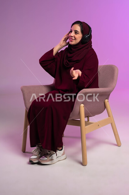 إستخدام تقنيات الهيدفون، بورتريه لسيدة عربية خليجية سعودية مبتسمة محجبة ترتدي العباءة الملونة تضع سماعات الرأس الصوتية تستمع إلى الموسيقى، إيماءات السعادة والاندماج، مفهوم الإصغاء والإستماع، الاستمتاع في أوقات الفراغ, خلفية بنفسجية