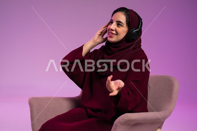 The concept of listening and hearing, the use of headphone techniques, a close-up portrait of a smiling Saudi Gulf Arab woman, veiled, wearing a colorful abaya, wearing audio headphones, listening to music, gestures of happiness and integration, enjoying leisure time, purple background