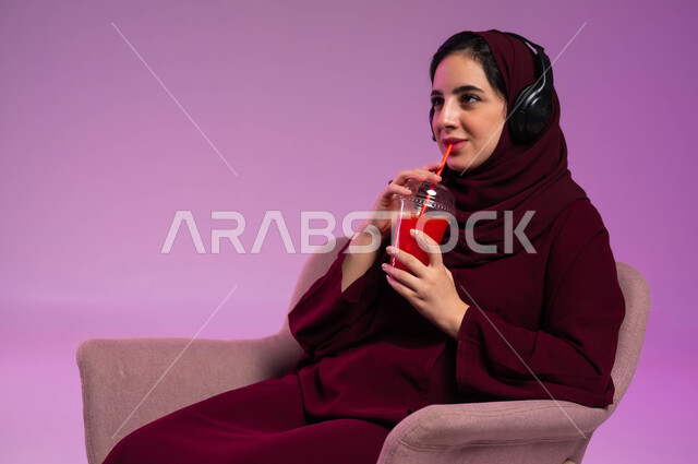 مشاهدة الأفلام والمسلسلات, عصائر طبيعية منعشة, إيماءات التفاعل والاندماج, بورتريه مقرب لفتاة عربية خليجية سعودية محجبة ترتدي العباءة الملونة وسماعة الرأس تجلس على كرسي مريح تقوم بشرب العصير, الاستمتاع في قضاء إجازة ممتعة, خلفية بنفسجية