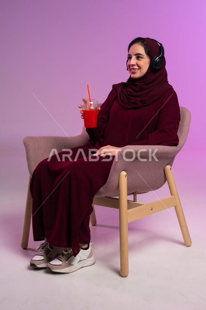 إيماءات الاندماج والتفاعل, عصائر طبيعية منعشة, بورتريه لفتاة عربية خليجية سعودية محجبة ترتدي العباءة الملونة وسماعة الرأس تجلس على كرسي مريح تمسك بيدها كأس العصير, الاستمتاع في قضاء إجازة ممتعة, مشاهدة الأفلام والمسلسلات, خلفية بنفسجية