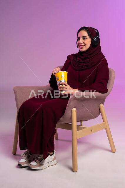 الاستمتاع بتناول البوشار، إيماءات التفاعل والاندماج, مفهوم الإصغاء والاستماع بورتريه لفتاة عربية خليجية سعودية محجبة ترتدي العباءة الملونة وسماعة الرأس تجلس على كرسي مريح, قضاء إجازة ممتعة, مشاهدة الأفلام والمسلسلات, خلفية بنفسجية
