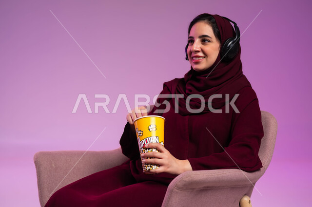 مشاهدة الأفلام والمسلسلات, إيماءات الفرح والسرور بورتريه مقرب لفتاة عربية خليجية سعودية محجبة ترتدي العباءة الملونة وسماعة الرأس تجلس على كرسي مريح تقوم بتناول البوشار, الاستمتاع في قضاء إجازة ممتعة, مفهوم الإصغاء والاستماع, خلفية بنفسجية