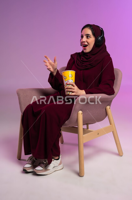 الاستمتاع في قضاء إجازة ممتعة, إيماءات الدهشة والذهول, بورتريه لفتاة عربية خليجية سعودية محجبة ترتدي العباءة الملونة وسماعة الرأس تجلس على كرسي مريح تقوم بتناول البوشار, مشاهدة الأفلام والمسلسلات, خلفية بنفسجية
