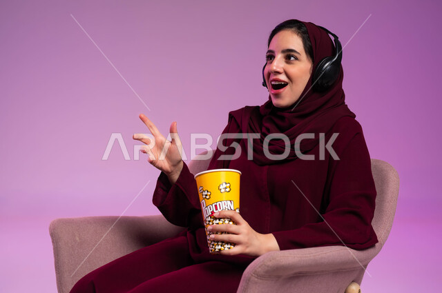 إيماءات الدهشة والذهول, بورتريه مقرب لفتاة عربية خليجية سعودية محجبة ترتدي العباءة الملونة وسماعة الرأس تجلس على كرسي مريح تقوم بتناول البوشار, الاستمتاع في قضاء إجازة ممتعة, مشاهدة الأفلام والمسلسلات, خلفية بنفسجية