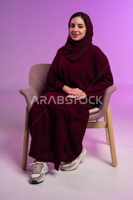 مفهوم الأنوثة والثقة بالنفس, بورتريه لفتاة عربية خليجية سعودية محجبة مبتسمة ترتدي العباءة الملونة تجلس على كرسي مريح تنظر إلى الكاميرا, الاهتمام بالأناقة والمظهر الخارجي, إيماءات السرور والفرح, خلفية بنفسجية