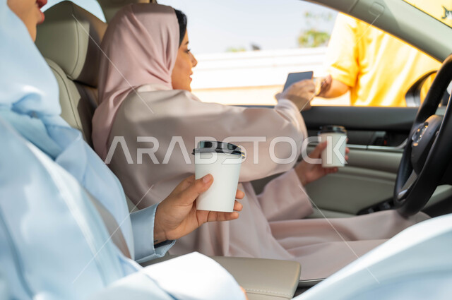 التنزه في أيام العطلة، مفهوم حرية القيادة للنساء السعوديات،استخدام الأجهزة التقنية الحديثه ،تناول المشروبات الساخنه مع الأصدقاء في الخارج ،امرأة عربيه خليجية سعودية محجبه برفقة صديقتها تقوم بالدفع الالكتروني من خلال الجوال عبر طلبات السيارة(الدرايف ثرو)