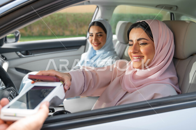 مفهوم الاعتماد على النفس في وسائل النقل ،استخدام الأجهزة التقنية الحديثه ،قضاء وقت ممتع مع الأصدقاء في الخارج ،حرية القيادة للنساء السعوديات، امرأة عربيه خليجية سعودية محجبه برفقة صديقتها تقوم بالدفع الالكتروني من خلال الجوال عبر طلبات السيارة(الدرايف ثرو)