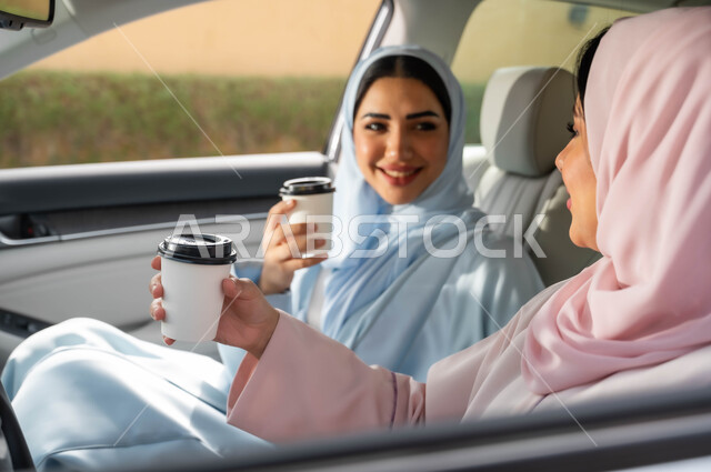 التجول في شوارع المملكة ،قضاء أوقات ممتعه مع الأصدقاء، حرية القيادة للنساء السعوديات، صورة مقربة لامرأة عربيه خليجية سعودية محجبه تجلس في مقعد السيارة برفقة صديقتها بإيماءات السرور، تناول المشروبات الساخنه، مفهوم الاستقلال و الاعتماد على النفس في وسائل النقل