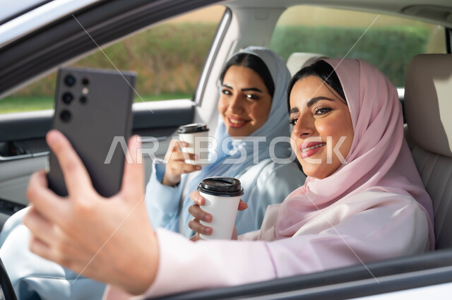تناول المشروبات الساخنه في الخارج ،توثيق اللحظات السعيدة عبر الجوال، مفهوم الترابط الأسري ،قضاء أوقات ممتعه مع الأخوة، التقاط صور السيلفي التذكارية عبر الجوال، صورة مقربة لامرأة عربية خليجية سعودية محجبه تحمل بيدها الهاتف المحمول تجلس في السيارة برفقة صديقتها