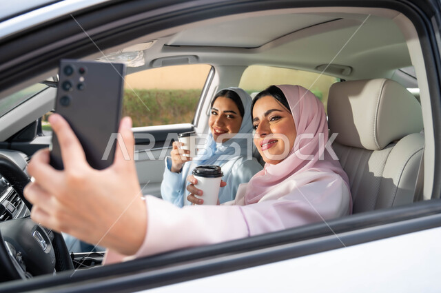 توثيق اللحظات السعيدة عبر الجوال، مفهوم الترابط الأسري ،قضاء أوقات ممتعه مع الأخوة، التقاط صور السيلفي التذكارية عبر الجوال، صورة مقربة لامرأة عربية خليجية سعودية محجبه تحمل بيدها الهاتف المحمول تجلس في السيارة برفقة صديقتها، تناول المشروبات الساخنه في الخارج