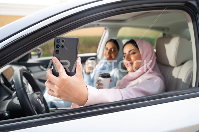 مفهوم الترابط الأسري ،قضاء أوقات ممتعه مع الأخوة، التقاط صور السيلفي التذكارية لتوثيق اللحظات السعيدة عبر الجوال، صورة مقربة لامرأة عربية خليجية سعودية محجبه تحمل بيدها الهاتف المحمول تجلس في السيارة برفقة صديقتها، تناول المشروبات الساخنه في الخارج