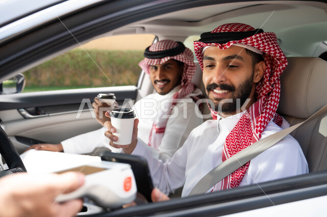 شراء المشروبات الساخنه في الخارج، الدفع الالكتروني من خلال الهاتف المحمول عبر طلبات (الدرايف ثرو)، استخدام التقنيات الحديثه المتطورة ،قضاء أوقات ممتعه مع الاصدقاء، رجلان عربيان خليجيان سعوديان يرتديان الزي التقليدي يجلسان في السيارة ،وسائل النقل والمواصلات