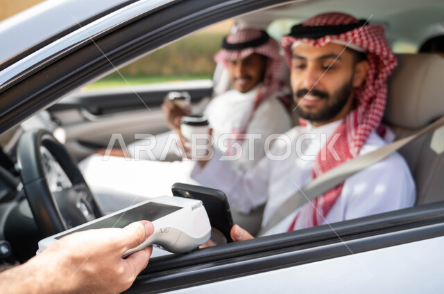 استخدام التقنيات الحديثه المتطورة ،قضاء أوقات ممتعه مع الاصدقاء، تناول المشروبات الساخنه، الدفع الالكتروني من خلال الهاتف المحمول عبر طلبات (الدرايف ثرو)، رجلان عربيان خليجيان سعوديان يرتديان الزي التقليدي يجلسان في السيارة ،وسائل النقل والمواصلات
