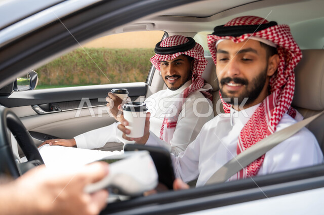 الدفع الالكتروني من خلال الهاتف المحمول عبر طلبات (الدرايف ثرو)، استخدام التقنيات الحديثه المتطورة ،قضاء أوقات ممتعه مع الاصدقاء، تناول المشروبات الساخنه، رجلان عربيان خليجيان سعوديان يرتديان الزي التقليدي يجلسان في السيارة ،وسائل النقل والمواصلات