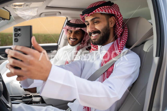 مفهوم الترابط العائلي ،قضاء أوقات ممتعه مع الأخوة، التقاط صور السيلفي التذكارية لتوثيق اللحظات السعيدة عبر الجوال، صورة مقربة لرجل عربي خليجي سعودي يرتدي الزي التقليدي ويحمل بيده الهاتف المحمول يجلس في السيارة برفقة صديقه بإيماءات السرور