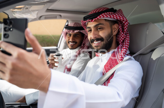 قضاء أوقات ممتعه مع الأخوة، التقاط صور السيلفي التذكارية لتوثيق اللحظات السعيدة عبر الجوال، صورة مقربة لرجل عربي خليجي سعودي يرتدي الزي التقليدي ويحمل بيده الهاتف المحمول يجلس في السيارة برفقة صديقه، تناول المشروبات الساخنه في الخارج