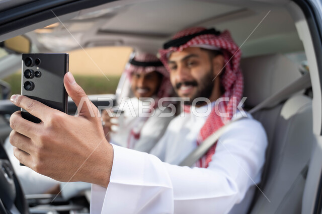 التقاط صور السيلفي التذكارية لتوثيق اللحظات السعيدة عبر الجوال، صورة مقربة ليد رجل عربي خليجي سعودي يرتدي الزي التقليدي ويحمل بيده الهاتف المحمول يجلس في السيارة برفقة صديقه، قضاء أوقات ممتعه مع الأخوة، تناول المشروبات الساخنه في الخارج