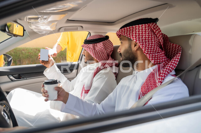 استخدام الأجهزة التقنية الحديثه ،قضاء أوقات ممتعه مع الاصدقاء، تناول المشروبات الساخنه، الدفع الالكتروني من خلال الهاتف المحمول عبر طلبات (الدرايف ثرو)، رجلان عربيان خليجيان سعوديان يرتديان الزي التقليدي يجلسان في السيارة بايماءات السرور ،وسائل النقل والمواصلات