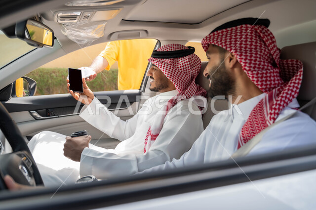 قضاء أوقات ممتعه مع الاصدقاء، تناول المشروبات الساخنه، الدفع الالكتروني من خلال الهاتف المحمول عبر طلبات (الدرايف ثرو)، رجلان عربيان خليجيان سعوديان يرتديان الزي التقليدي يجلسان في السيارة بايماءات السرور ،استخدام الأجهزة التقنية الحديثه ،وسائل النقل والمواصلات