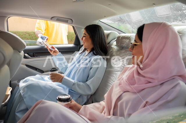 Enjoying walking in the streets of the Kingdom, drinking hot drinks, electronic payment through the mobile phone via drive-thru orders, a veiled Saudi Arabian Gulf woman sitting in the car seat with her friend, using modern technical devices, means of transport and transportation.