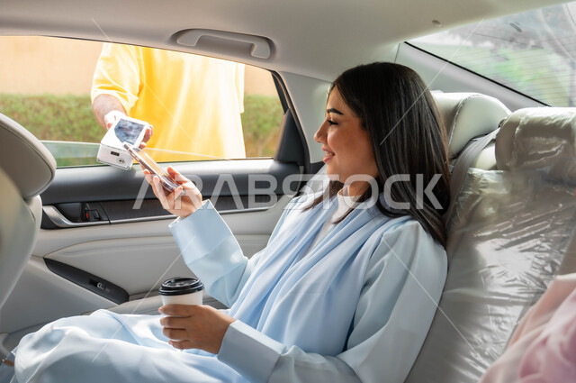 تناول المشروبات الساخنه، الدفع الالكتروني من خلال الهاتف المحمول عبر طلبات (الدرايف ثرو)، امرأة عربيه خليجية سعودية محجبه تجلس في مقعد السيارة تقوم باستخدام الأجهزة التقنية الحديثه ،وسائل النقل والمواصلات، التنزه في شوارع المملكة