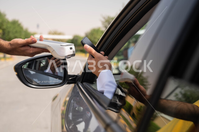 دمج التكنولوجيا في الحياة اليومية لتوفير راحة أكبر للأفراد ،الدفع الالكتروني من خلال الهاتف المحمول عبر طلبات السيارة(الدرايف ثرو)، صورة مقربة ليد عربي خليجي سعودي يقوم باستخدام الأجهزة التقنية الحديثه ،التنقل في شوارع المملكة