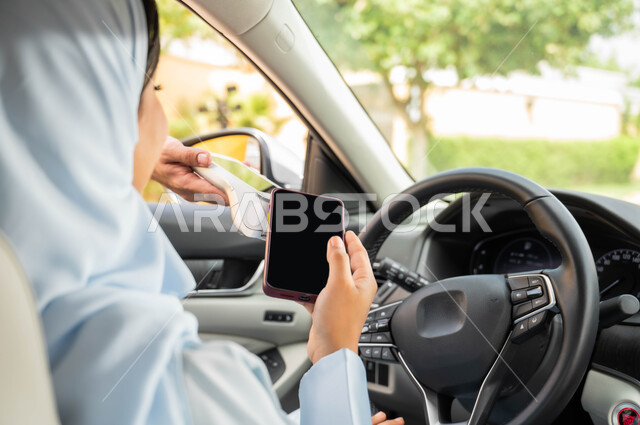 الأجهزة التقنية الحديثه الالكترونية ،حرية القيادة للنساء السعوديات، صورة مقربة من الخلف لامرأة عربيه خليجية سعودية محجبه تقوم بالدفع الالكتروني من خلال الهاتف المحمول عبر طلبات السيارة(الدرايف ثرو)، مفهوم الاستقلال و الاعتماد على النفس في وسائل النقل والمواصلات